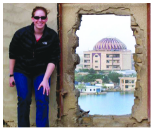 Image of a person standing next to a hole in the wall reaving a building in the background. 